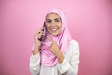 Genç, güzel bir Arap kadın, izole edilmiş pembe arka planda tesettürlü, İslami başörtüsü takıyor. Gülümsüyor, kulağındaki telefona bakıyor.