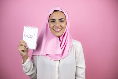 Genç, güzel, Arap bir kadın, soyutlanmış pembe arka plan üzerine tesettür takıyor. Korktu ve şaşırdı. Elinde not defteri tutarken tek kelime etmedi.