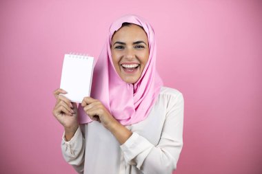 Genç, güzel bir Arap kadın, izole edilmiş pembe arka planda İslami başörtüsü takıyor, gülümsüyor ve elinde boş bir defter gösteriyor.