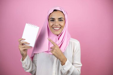 Genç, güzel bir Arap kadın, izole edilmiş pembe arka planda İslami başörtüsü takıyor, gülümsüyor ve elinde boş bir defter gösteriyor.