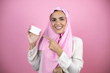 Genç, güzel bir Arap kadın, izole edilmiş pembe arka planda İslami başörtüsü takıyor gülümsüyor ve elinde beyaz bir kart tutuyor.