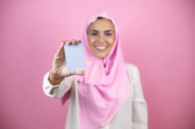Genç, güzel bir Arap kadın, izole edilmiş pembe arka planda İslami başörtüsü takıyor gülümsüyor ve elinde beyaz bir kart tutuyor.