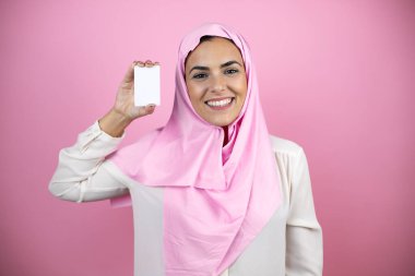 Genç, güzel bir Arap kadın, izole edilmiş pembe arka planda İslami başörtüsü takıyor gülümsüyor ve elinde beyaz bir kart tutuyor.