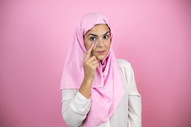 Genç, güzel bir Arap kadın, izole edilmiş pembe arka plan üzerine tesettür takmış. Gözlerine işaret ediyor. Hareketlerini izliyor.