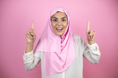 Genç, güzel bir Arap kadın soyutlanmış pembe arka planda tesettür takıyor gülümsüyor, kameraya bakıyor ve parmaklarıyla işaret ediyor ve kollarını kaldırıyor.