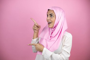 İzole edilmiş pembe arka plan üzerinde İslami başörtüsü takan genç, güzel bir Arap kadın şaşırmış ve parmaklarını işaret ediyor.