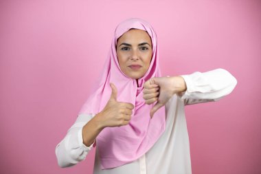 Genç, güzel bir Arap kadın, izole edilmiş pembe arka plan üzerinde başörtüsü takıyor. Baş parmaklarını yukarı aşağı hareket ettiriyor, anlaşmazlık ve anlaşma ifadesi takınıyor. Çılgın çatışma