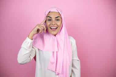 Genç, güzel bir Arap kadın, izole edilmiş pembe arka plan üzerine tesettür takıyor, gülümsüyor ve parmakları kafasında bir fikri olduğunu düşünüyor..