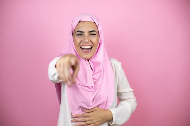 Genç, güzel bir Arap kadın, izole edilmiş pembe arka plan üzerine tesettür takmış sana gülüyor, eli vücudunun üzerinde kamerayı işaret ediyor, utanç ifadesi.