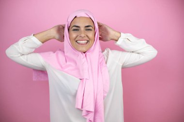 Genç, güzel bir Arap kadın, izole edilmiş pembe arka planda tesettür takıyor. Rahatlıyor ve esniyor, kolları ve elleri başının ve boynunun arkasında, gülümsüyor.