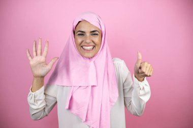 Genç, güzel bir Arap kadın, izole edilmiş pembe arka planda İslami başörtüsü takıyor. Altıncı parmaklarıyla işaret ederken, kendinden emin ve mutlu gülümsüyor.