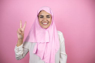 Genç, güzel bir Arap kadın, izole edilmiş pembe arka planda İslami başörtüsü takıyor ve parmaklarıyla iki numarayı gösterirken kendinden emin ve mutlu bir şekilde gülümsüyor.