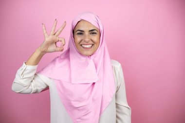 İzole edilmiş pembe arka planda İslami tesettür takan genç, güzel bir Arap kadın tamam işareti yapıyor parmaklarıyla ve gülümsüyor, mükemmel bir sembol.