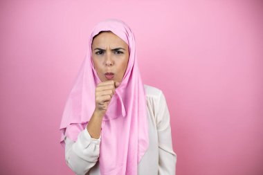 Genç, güzel bir Arap kadın, izole edilmiş pembe arka planda İslami başörtüsü takıyor. Elleri ağzına dayanıyor çünkü öksürüyor.