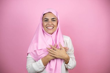 Genç, güzel bir Arap kadın soyutlanmış pembe arka planda tesettür takıyor. Elleri göğsünde ve yüzünde minnet dolu bir jest ile gülümsüyor..