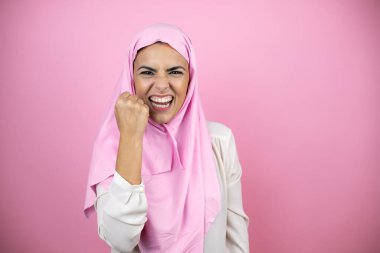 Genç, güzel bir Arap kadın, izole edilmiş pembe arka plan üzerine tesettür takıyor kızgın ve kızgın yumruğunu kaldırıyor ve öfkeyle bağırıyor. Öfke ve agresif kavram.
