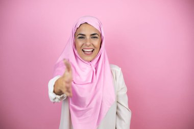İzole edilmiş pembe arka planda İslami başörtüsü takan genç, güzel bir Arap kadın selamlama ve hoş geldin demek için gülümseyerek tokalaşıyor.