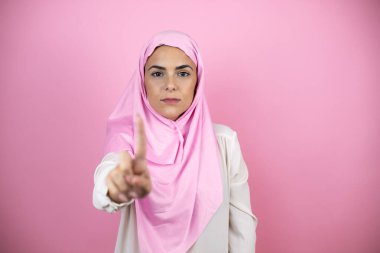 İzole edilmiş pembe arka plan üzerinde İslami başörtüsü takan genç ve güzel bir Arap kadın bir numaralı parmaklarıyla işaret ediyor.