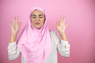 Genç, güzel bir Arap kadın, izole edilmiş pembe arka planda İslami başörtüsü takıyor. Rahatlayın ve gözleriniz kapalı gülümseyin. Parmaklarınızla meditasyon hareketi yapın.