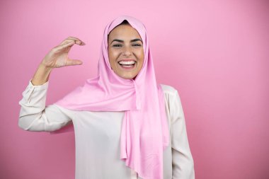 Genç, güzel bir Arap kadın, izole edilmiş pembe arka planda tesettür takıyor, gülümsüyor ve elleriyle küçük boy işareti yapıyor. Ölçü kavramı.