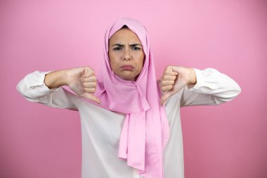 Genç, güzel bir Arap kadın, izole edilmiş pembe arka planda başörtüsü takıyor. Kızgın bir yüzü var.
