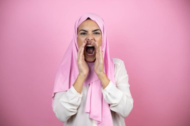 Genç, güzel bir Arap kadın, izole edilmiş pembe arka planda İslami başörtüsü takıyor. Elleri ağızda bağırarak ve yüksek sesle bağırıyor.