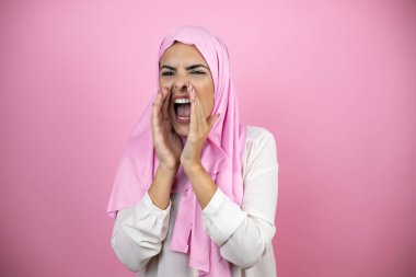 Genç, güzel bir Arap kadın, izole edilmiş pembe arka planda İslami başörtüsü takıyor. Elleri ağızda bağırarak ve yüksek sesle bağırıyor.