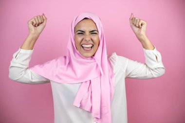 Genç, güzel bir Arap kadın, izole edilmiş pembe arka plan üzerine tesettür takıyor. Çok mutlu ve heyecanlı. Kollarını kaldırıp, gülümseyerek ve başarı için bağırarak, kazanan jestler yapıyor..
