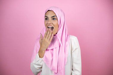 Genç, güzel bir Arap kadın, soyutlanmış pembe arka plan üzerine tesettüre bürünmüş, eli ağzında ve şaşırmış, yan bakıyor.