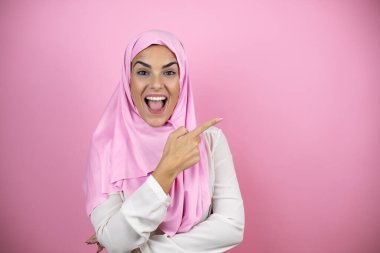Genç, güzel bir Arap kadın, izole edilmiş pembe arka plan üzerine tesettür takıyor. Gülümsüyor ve parmaklarını yana doğru uzatıyor.