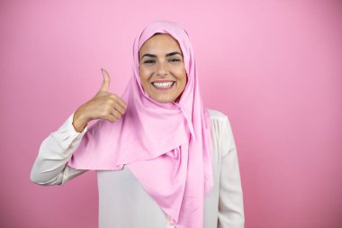 Genç, güzel bir Arap kadın, izole edilmiş pembe arka planda İslami başörtüsü takıyor. Gülümsüyor ve baş parmağıyla işaret veriyor.