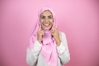İzole edilmiş pembe arka planda İslami başörtüsü takan genç, güzel bir Arap kadın gülümsüyor ve parmaklarıyla ve ağzıyla işaret ediyor.