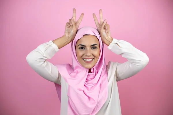 若いです美しいですArab女性身に着けていますイスラムヒジャーブ上の隔離されたピンクの背景面白いとクレイジーをポーズ指で頭の上にウサギの耳 笑顔陽気に — ストック写真
