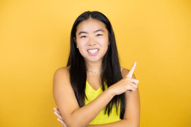 Sarı arka plan üzerinde genç ve güzel Asyalı bir kadın. Gülümsüyor ve parmaklarını yana doğru uzatıyor.