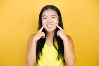 Sarı arka plan üzerinde genç ve güzel Asyalı bir kadın. Gülümsüyor ve parmaklarıyla ve ağzıyla işaret ediyor.