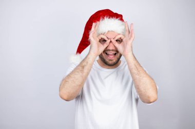 Beyaz arka planda Noel Baba şapkası takan genç yakışıklı adam gülümseyen yüzüyle, parmaklarından bakan gözleriyle şaşırmış bir jest yapıyor.