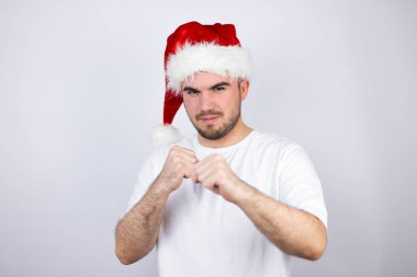 Beyaz arka planda Noel Baba şapkası takan genç yakışıklı adam dövüşmek için yumruk, agresif ve öfkeli saldırı, tehdit ve şiddet.