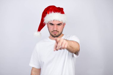 Beyaz arka planda Noel Baba şapkası takan genç ve yakışıklı bir adam ön tarafı işaret ediyor.