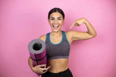 İzole edilmiş pembe arka planda spor kıyafetleri giyen genç ve güzel bir kadın. Kendine güvenen, yüzünde bir gülümsemeyle kendini gururlu ve mutlu olarak gösteren bir kadın..