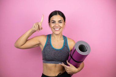 İzole edilmiş pembe arka planda spor giyimli genç bir kadın gülümsüyor, kameraya bakıyor, parmaklarıyla işaret ediyor ve kollarını kaldırıyor. Elinde kıymık tutuyordu..