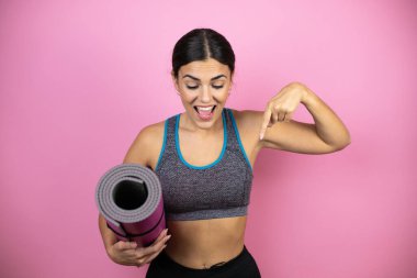 İzole edilmiş pembe arka planda spor giyimli genç ve güzel bir kadın şaşırmış bir şekilde parmağını işaret ediyor..