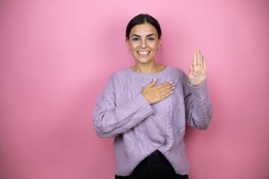 Güzel bir kadın pembe arka planda sıradan bir mor kazak giyiyor gülümsüyor, elleri göğsünde ve parmakları havada küfür ediyor, sadakat yemini ediyor.