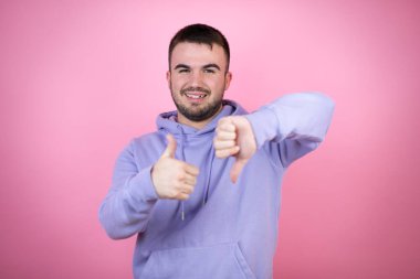 Soyutlanmış pembe arka plan üzerine sıradan bir eşofman giyen genç yakışıklı adam baş parmak kaldırıyor, anlaşmazlık ve anlaşma ifadesi sergiliyor. Çılgın çatışma