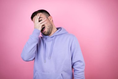 İzole edilmiş pembe arka plan üzerinde sıradan bir eşofman üstü giyen genç yakışıklı adam şok içinde yüzünü ve gözlerini kapayarak utangaç ifadesiyle parmaklarının arasından bakıyor.