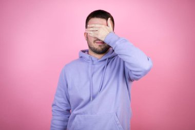 İzole edilmiş pembe arka plan üzerine gündelik kazak giyen genç yakışıklı adam ciddi ve eliyle gözlerini kapatıyor.