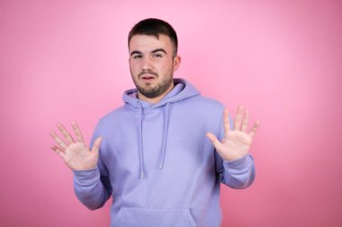 Soyutlanmış pembe arka plan üzerinde sıradan bir eşofman üstü giyen genç yakışıklı adam korku ifadesinden korkuyor ve dehşete düşüyor. El hareketlerini bırak, şok içinde bağır. Panik konsepti