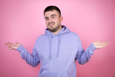 İzole edilmiş pembe arka plan üzerine sıradan eşofman üstü giyen genç yakışıklı bir adam. Kolları ve elleri havada, kafası karışmış bir ifade.