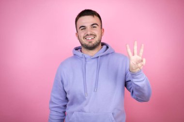 İzole edilmiş pembe arka planda sıradan bir eşofman üstü giyen genç yakışıklı adam üç numaralı parmaklarıyla gülümseyerek kendinden emin ve mutlu bir şekilde onu işaret ediyor.