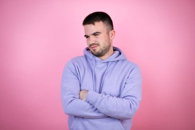 Soyutlanmış pembe arka plan üzerine sıradan bir eşofman giyen genç yakışıklı yorgun ve kollarını kavuşturmaktan sıkılmış görünüyor.