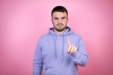 Soyutlanmış pembe arka planda sıradan bir eşofman üstü giyen genç yakışıklı bir numaralı parmaklarıyla işaret etmek ciddidir.